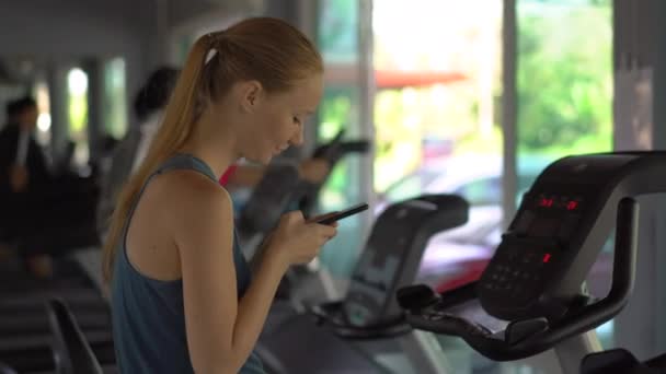 Uma jovem mulher no ginásio em uma bicicleta de exercício segurando o telefone na frente de seus olhos em suas mãos. Toda a atenção está no telefone. O conceito de dependência das redes sociais. Dependência móvel — Vídeo de Stock