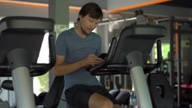 Telefonda onun gözlerinin önünde onun elinde tutan bir egzersiz bisikleti üzerinde spor salonunda genç bir adam. Tüm dikkat telefon olduğunu. Sosyal ağlar üzerinde bağımlılık kavramı. Mobil bağımlılığı