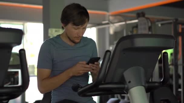 En ung man på gymmet på en motionscykel som håller telefonen framför hans ögon i hans händer. All uppmärksamhet är i telefonen. Begreppet beroende på sociala nätverk. Mobila missbruk — Stockvideo