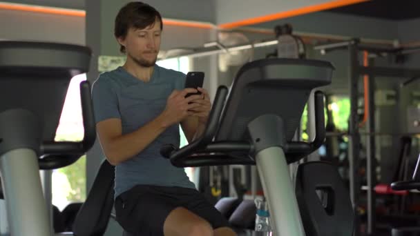 Молодий чоловік у спортзалі на тренажерному велосипеді тримає телефон перед очима в руках. Вся увага в телефоні. Поняття залежності від соціальних мереж. Мобільна залежність — стокове відео