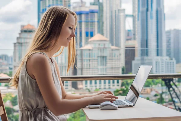 Jonge vrouw is bezig met een laptop op haar balkon met uitzicht op de wolkenkrabbers. Freelancer, externe werkzaamheden, werken vanuit huis — Stockfoto
