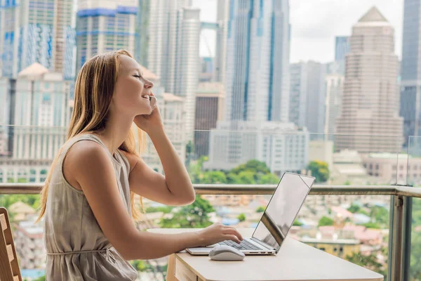 Jonge vrouw is bezig met een laptop op haar balkon met uitzicht op de wolkenkrabbers. Freelancer, externe werkzaamheden, werken vanuit huis — Stockfoto