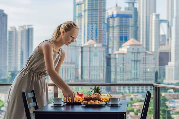 Молодая женщина лежит на столе. Стол для завтрака с фруктами и круассаном на балконе на фоне большого города — стоковое фото