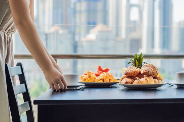Mladá žena leží na stole. Snídani s kávou ovoce a chléb croisant na balkóně na pozadí velkoměsta — Stock fotografie