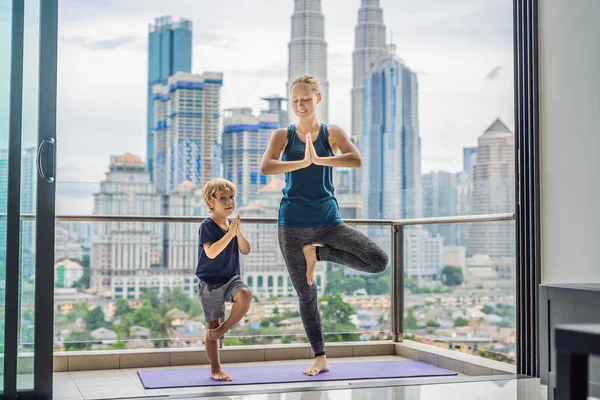 Mom and son are practicing yoga on the balcony in the background of a big city. Sports mom with kid doing morning work-out at home. Mum and child do the exercises together, healthy family lifestyle