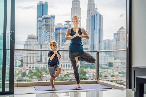 Moeder en zoon zijn het beoefenen van yoga op het balkon op de achtergrond van een grote stad. Sporten moeder met kind doen ochtend trainen thuis. Mama en kind doen de oefeningen samen, gezonde familie leefstijl — Stockfoto