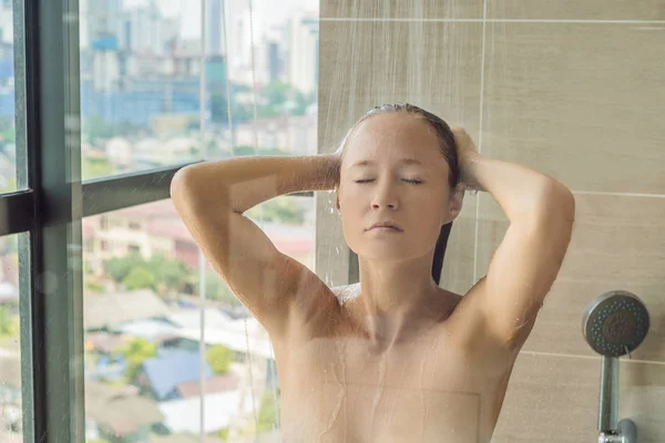 Mooie vrouw in de douche achter glas met druppels op de achtergrond van een venster met een panoramisch uitzicht over de stad — Stockfoto