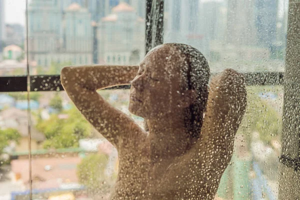 Красивая женщина в душе за стеклом с капельками на фоне окна с панорамным видом на город — стоковое фото