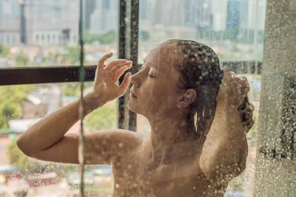 Schöne Frau in der Dusche hinter Glas mit Tropfen auf dem Hintergrund eines Fensters mit einem Panoramablick auf die Stadt — Stockfoto