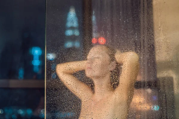 Hermosa mujer en la ducha detrás de vidrio con gotas en el fondo de una ventana con una vista panorámica de la ciudad nocturna — Foto de Stock