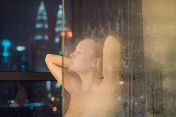 Mulher bonita no chuveiro atrás de vidro com gotas no fundo de uma janela com uma vista panorâmica da cidade noturna — Fotografia de Stock