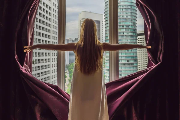 Young woman opens the window curtains and looks at the skyscrapers in the big city