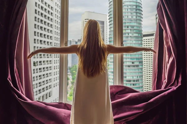 Молода жінка відкриває віконні штори і дивиться на хмарочоси у великому місті — стокове фото