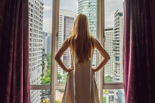 Mujer joven abre las cortinas de la ventana y mira a los rascacielos en la gran ciudad — Foto de Stock