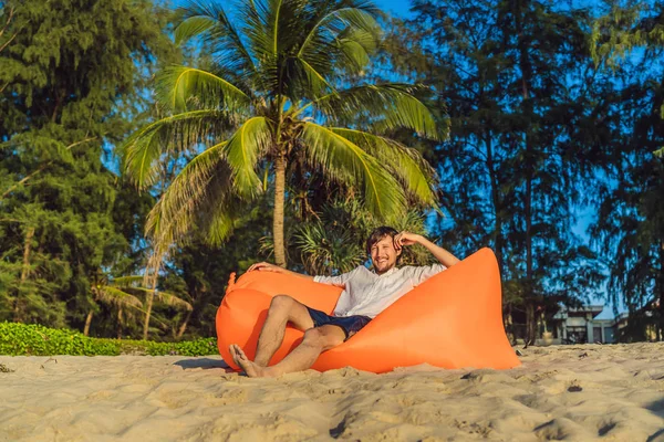 Retrato de estilo de vida de verano del hombre sentado en el sofá inflable naranja en la playa de la isla tropical. Relajarse y disfrutar de la vida en la cama de aire —  Fotos de Stock