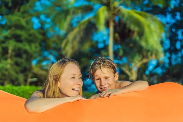 Summer lifestyle portrait of mother and son sitting on the orange inflatable sofa on the beach of tropical island. Relaxing and enjoying life on air bed