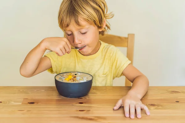 Niño feliz sentado y comiendo un tazón de batido fresco con mango, fruta de dragón, granola y semillas de chía — Foto de Stock