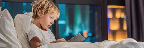 Мальчик использует скрижаль в своей постели, прежде чем заснуть на фоне ночного города. Детские и технологические концепции BANNER, длинный формат — стоковое фото