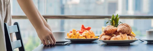 A jovem está deitada numa mesa. Mesa de pequeno-almoço com frutas de café e pão croisant em uma varanda contra o pano de fundo da cidade grande BANNER, LONG FORMAT — Fotografia de Stock