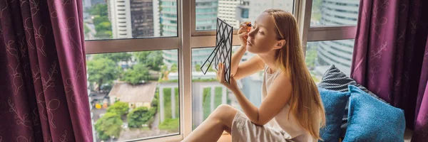 Junge Frau schminkt sich am Fenster sitzend mit Blick auf die Wolkenkratzer und das Großstadtbanner, Langformat — Stockfoto