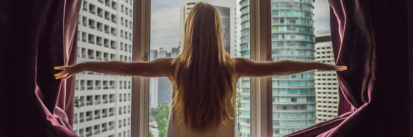 Young woman opens the window curtains and looks at the skyscrapers in the big city BANNER, LONG FORMAT