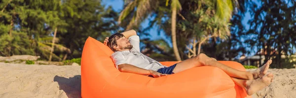 Retrato de estilo de vida de verano del hombre sentado en el sofá inflable naranja en la playa de la isla tropical. Relajarse y disfrutar de la vida en la cama de aire BANNER, FORMATO LARGO — Foto de Stock