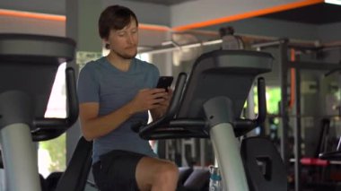 Telefonda onun gözlerinin önünde onun elinde tutan bir egzersiz bisikleti üzerinde spor salonunda genç bir adam. Tüm dikkat telefon olduğunu. Sosyal ağlar üzerinde bağımlılık kavramı. Mobil bağımlılığı