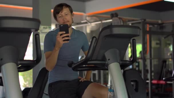 En ung man på gymmet på en motionscykel som håller telefonen framför hans ögon i hans händer. All uppmärksamhet är i telefonen. Begreppet beroende på sociala nätverk. Mobila missbruk — Stockvideo