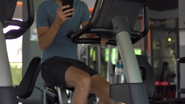 Um jovem no ginásio em uma bicicleta de exercício segurando o telefone na frente de seus olhos em suas mãos. Toda a atenção está no telefone. O conceito de dependência das redes sociais. Dependência móvel — Vídeo de Stock