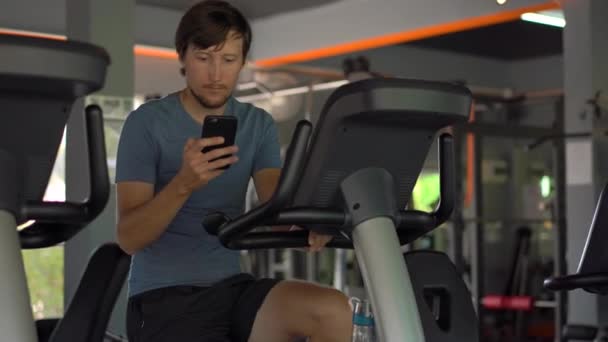 En ung man på gymmet på en motionscykel som håller telefonen framför hans ögon i hans händer. All uppmärksamhet är i telefonen. Begreppet beroende på sociala nätverk. Mobila missbruk — Stockvideo