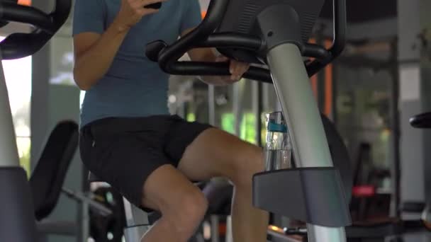 Um jovem no ginásio em uma bicicleta de exercício segurando o telefone na frente de seus olhos em suas mãos. Toda a atenção está no telefone. O conceito de dependência das redes sociais. Dependência móvel — Vídeo de Stock