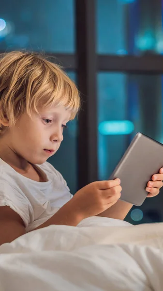 Chlapec používá tabletu v posteli, než spí na pozadí nočního města. Koncepty pro děti a technologie svislý formát pro článek Instagram mobilní text nebo velikost příběhů. Mobilní Tapeta — Stock fotografie