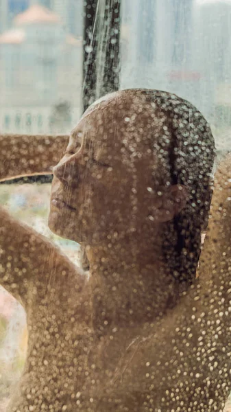 Mooie vrouw in de douche achter glas met druppels op de achtergrond van een venster met een panoramisch uitzicht over de stad verticale indeling voor mobiele verhaal Instagram of verhalen grootte. Mobiele achtergronden — Stockfoto