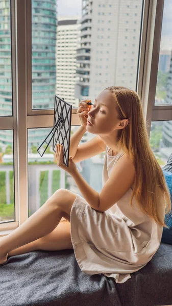 Mujer joven hace maquillaje sentado junto a la ventana con una vista panorámica de los rascacielos y la gran ciudad FORMATO VERTICAL para Instagram historia móvil o el tamaño de las historias. Fondo de pantalla móvil — Foto de Stock