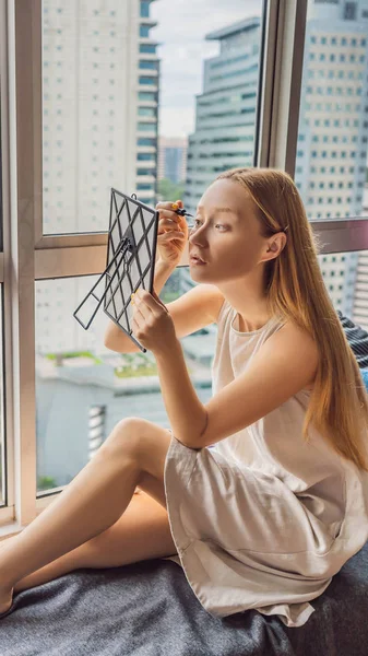 Молодая женщина делает макияж сидя у окна с панорамным видом на небоскребы и большой город ВЕРТИЧЕСКИЙ ФОРМАТ для мобильной истории Instagram или размер историй. Мобильные обои — стоковое фото
