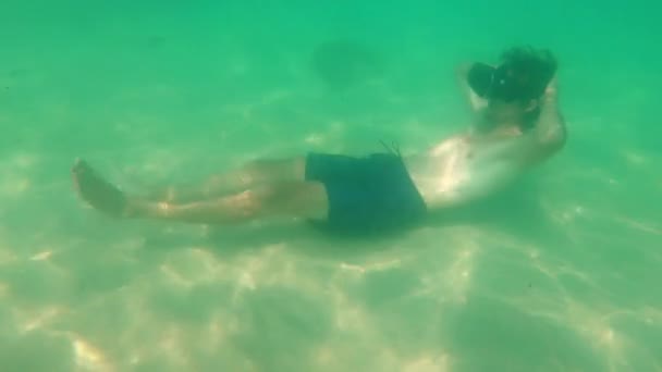 Underwater shot of a young man use a VR headset laying on a sea bottom. A person wearing a vr glasses feels like he is ina tropical sea. Augmented reality concept — Stock Video