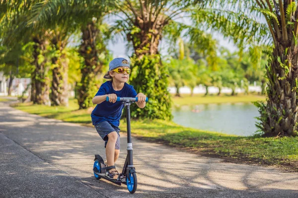 Bambino su scooter calcio nel parco. I bambini imparano a pattinare a rotelle. Ragazzino che pattina nella soleggiata giornata estiva. Attività all'aperto per bambini su strada residenziale sicura. Sport attivo per bambini in età prescolare — Foto Stock