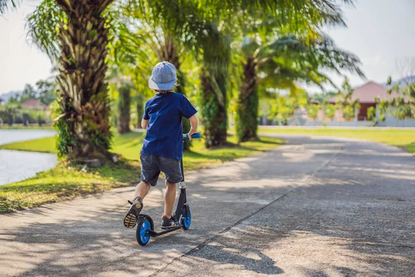 Bambino su scooter calcio nel parco. I bambini imparano a pattinare a rotelle. Ragazzino che pattina nella soleggiata giornata estiva. Attività all'aperto per bambini su strada residenziale sicura. Sport attivo per bambini in età prescolare — Foto Stock