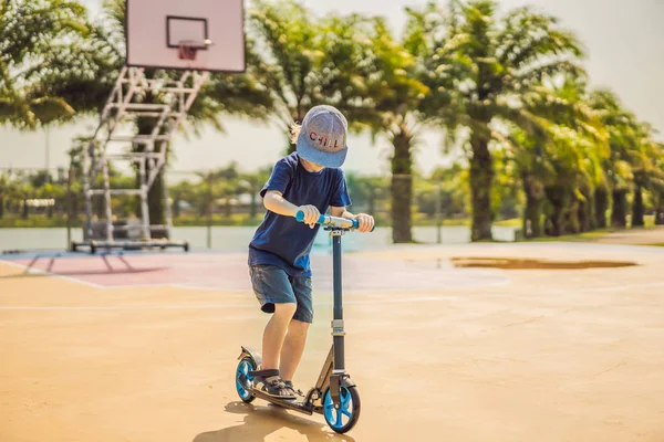 농구 코트에 킥 스쿠터에 행복한 아이. 아이들은 롤러 보드를 스케이트하는 법을 배웁니다. 화창한 여름 날에 스케이트를 타는 어린 소년. 안전한 주거 거리에아이들을위한 야외 활동. 액티브 스포츠 — 스톡 사진