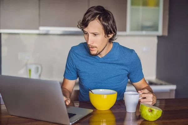 Молодой человек завтракает и пользуется ноутбуком на кухне — стоковое фото
