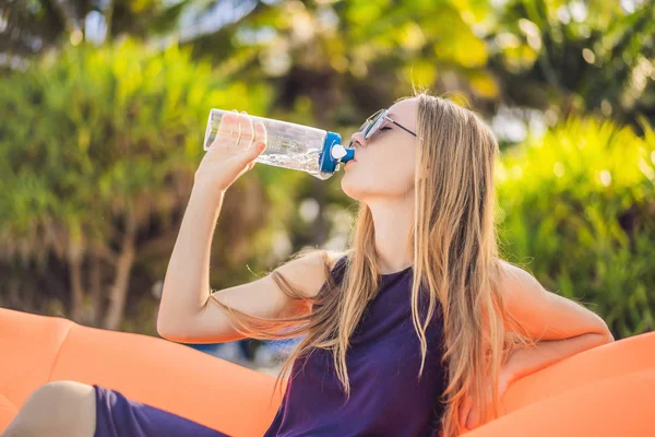 Summer lifestyle portrait of pretty girl sitting on the orange inflatable sofa and drinking water on the beach of tropical island. Relaxing and enjoying life on air bed — Stock Photo, Image