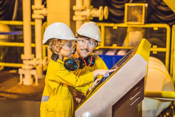 Mladý muž a malý chlapec jsou ve žluté pracovní uniformě, skleničkách a helmici v průmyslovém prostředí, olejová plošina nebo zařízení na zkapalněný plyn, které hledí na obrazovku — Stock fotografie