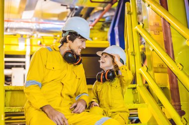 Young man and a little boy are both in a yellow work uniform, glasses, and helmet in an industrial environment, oil Platform or liquefied gas plant