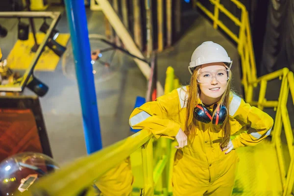 Giovane donna in uniforme da lavoro gialla, occhiali e casco in ambiente industriale, piattaforma petrolifera o impianto a gas liquefatto — Foto Stock