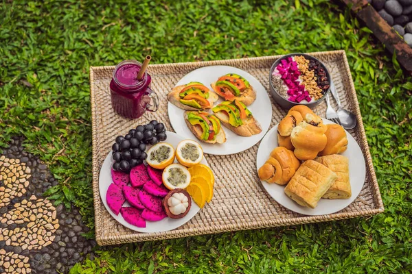 과일, 빵, 아보카도 샌드위치, 잔디 위에 서있는 스무디 그릇과 트레이에 아침 식사 — 스톡 사진