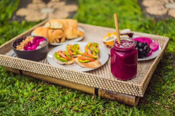 Café da manhã em uma bandeja com frutas, pães, sanduíches de abacate, tigela de smoothie em pé na grama — Fotografia de Stock