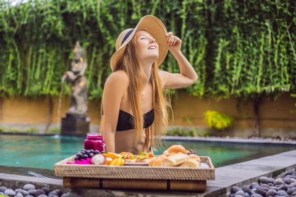 Young woman eating breakfast on a tray with fruit, buns, avocado sandwiches, smoothie bowl by the pool. Summer healthy diet, vegan breakfast. Tasty vacation concept