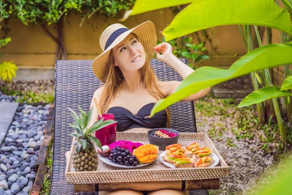 Young woman eating breakfast in a lounge chair on a tray with fruit, buns, avocado sandwiches, smoothie bowl by the pool. Summer healthy diet, vegan breakfast. Tasty vacation concept
