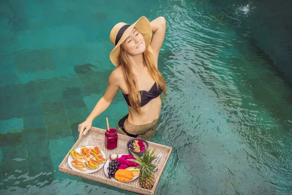 Breakfast tray in swimming pool, floating breakfast in luxury hotel. Girl relaxing in the pool drinking smoothies and eating fruit plate, smoothie bowl by the hotel pool. Exotic summer diet. Tropical