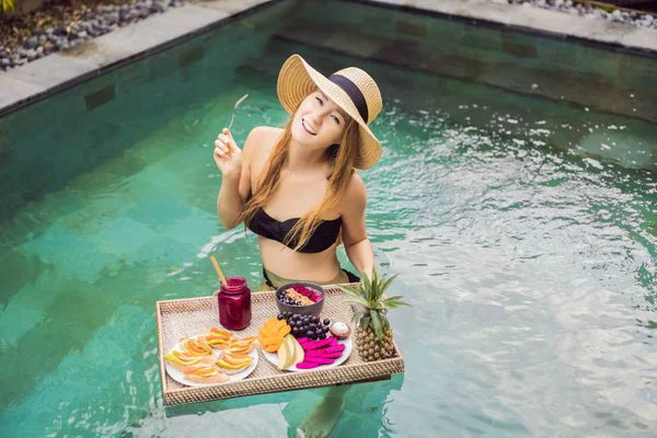 Breakfast tray in swimming pool, floating breakfast in luxury hotel. Girl relaxing in the pool drinking smoothies and eating fruit plate, smoothie bowl by the hotel pool. Exotic summer diet. Tropical